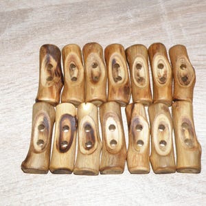 Puede incluir: Una colección de doce botones de madera tipo palanca. Cada botón es de color marrón claro con un patrón de vetas de madera natural. Los botones tienen dos agujeros para coser y están dispuestos en forma rectangular.