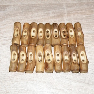 Pode incluir: Uma coleção de botões de madeira com uma cor castanha clara natural. Cada botão é cilíndrico com dois pares de orifícios para costura. Os botões têm uma aparência rústica e orgânica, sugerindo que são feitos de um material natural.