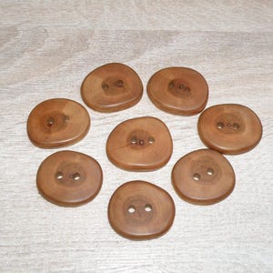Puede incluir: Una colección de ocho botones de madera redondos de color marrón claro dispuestos en un patrón circular. Cada botón presenta una superficie lisa y pulida con patrones de veta de madera natural visibles y dos pequeños agujeros en el centro.