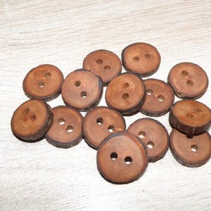Pode incluir: Um conjunto de catorze botões circulares de madeira, cada um com uma borda de casca natural e dois orifícios centrais. Os botões exibem uma superfície castanha lisa e polida com padrões de grão de madeira visíveis e formas circulares variadas.