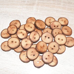 Puede incluir: Una colección de botones de madera redondos de color marrón claro, cada uno con dos orificios centrales. Los botones tienen un borde natural e irregular con textura de corteza visible, mostrando variados patrones y tonos de madera.
