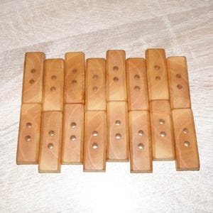 Pode incluir: Trivet de madeira composto por blocos retangulares. Cada bloco é castanho claro com dois rebites de metal. O trivet foi projetado para proteger as superfícies de pratos quentes e está disposto em um padrão de grade.