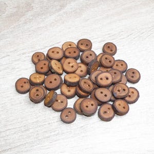 Peut inclure: Une collection de petits boutons ronds en bois brun. Chaque bouton présente une surface lisse et plate avec deux trous centraux pour la fixation et un bord texturé brun foncé rappelant l'écorce, mettant en valeur le grain naturel du bois.