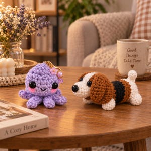 May include: A lavender crocheted octopus keychain with black eyes and pink cheeks alongside a small crocheted beagle dog figurine featuring brown, black, and white yarn, both displayed on a wooden surface near a mug with the text Good Things Take Time.