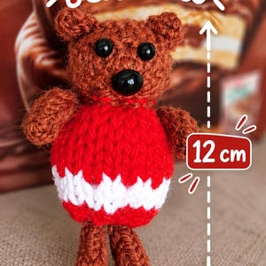 May include: A brown crocheted bear toy with black bead eyes and a black nose, wearing a red sweater with a white horizontal stripe. The bear is 12 cm tall, as indicated by a measurement graphic. The text Knitted Pattern is visible above.