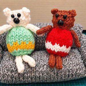 May include: Two small, hand-knitted stuffed animals, one white and one brown, sitting on a miniature grey knitted sofa. The white animal wears a light green and yellow sweater, while the brown animal wears a red and white sweater. The text PDF is visible.
