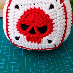 Puede incluir: Un cubo de ganchillo con base blanca y detalles en rojo y negro. Cada cara muestra un motivo de calavera roja centrado en un cuadrado blanco, enmarcado por bordes de hilo rojo. El cubo está colocado sobre una base de corte verde.