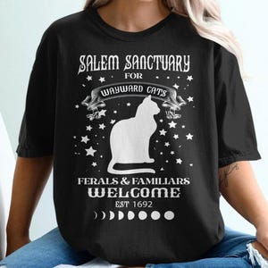 May include: Black t-shirt with white text that reads "Salem Sanctuary for Wayward Cats, Ferals & Familiars Welcome, Est 1692". The shirt also features a white silhouette of a cat and stars.