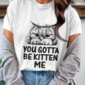 May include: White t-shirt with a black and white illustration of a cat peeking over the top. The text "You gotta be kitten me" is printed below the cat.