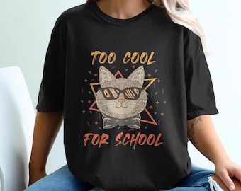 May include: Black t-shirt with a graphic of a cat wearing sunglasses and a bow tie. The text "Too Cool For School" is printed on the shirt.