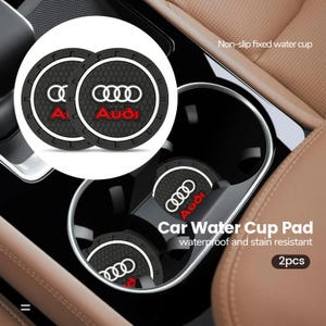 May include: Two circular black rubber car cup holder coasters featuring a textured surface, the white Audi four-ring logo, and the red Audi brand name. These non-slip, waterproof, and stain-resistant pads are designed to fit into vehicle cup holders.