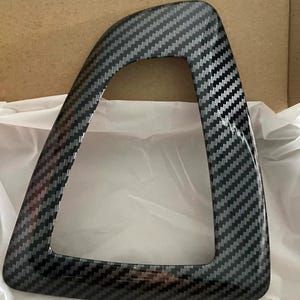 May include: A black carbon fibre trim piece with a rectangular opening. The trim piece has a textured surface and is designed to fit around a car's interior.