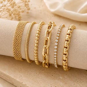 May include: A set of six gold-toned bracelets displayed on a cylindrical stand. The collection includes a wide mesh band, a smooth snake chain, a beaded chain, a thick oval link chain, a delicate crystal-encrusted tennis chain, and a box link chain.