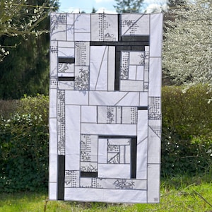 Op de afbeelding: Een wit quilt met een geometrisch patroon van zwarte en grijze vierkanten en rechthoeken. Het quilt hangt aan een waslijn buiten.