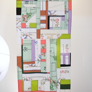 Op de afbeelding: Een patchworkdeken gemaakt van verschillende stoffen met bloemenpatronen en geometrische ontwerpen in tinten groen, roze, wit en bruin. De deken hangt aan een witte muur.