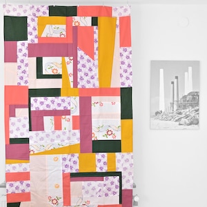 Op de afbeelding: Een kleurrijke patchwork quilt met een geometrisch ontwerp. De quilt heeft rechthoekige en vierkante blokken in tinten roze, koraal, geel, groen en wit, met bloemen- en patroonstoffen. Een zwart-wit foto van industriële structuren hangt aan de muur.