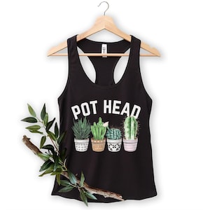Può includere: Una canotta nera a vogatore con la scritta bianca POT HEAD sopra un grafico raffigurante quattro distinti cactus verdi in vasi con vari motivi. Il capo è esposto su una gruccia di legno con un ramo decorativo di foglie appoggiato davanti.