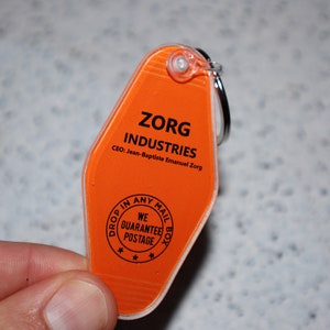Op de afbeelding: Oranje plastic motel sleutelhanger met de tekst "ZORG INDUSTRIES" en "CEO: Jean-Baptiste Emanuel Zorg". De achterkant van de sleutelhanger heeft een cirkel met de tekst "DROP IN ANY MAIL BOX WE GUARANTEE POSTAGE" en drie sterren.