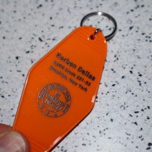 Op de afbeelding: Oranje plastic hotel sleutelhanger met de tekst "Korben Dallas 5,000 block 287-53 Brooklyn, New York" en een zilveren cirkel met de tekst "Drop In Any Time We Guarantee  Parking".
