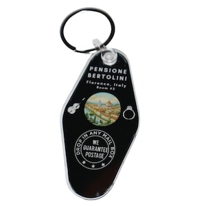 Op de afbeelding: Zwarte en witte plastic sleutelhanger in de vorm van een motel-sleutel met de tekst "PENSIONE BERTOLINI Florence, Italië Kamer #3" en een klein beeld van een stadsgezicht. De sleutelhanger heeft ook de tekst "DROP IN ANY MAIL BOX WE GUARANTEE POSTAGE" in een cirkel.