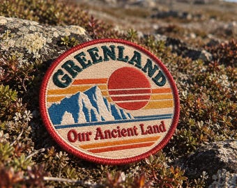 May include: A circular embroidered patch featuring a landscape design with a blue mountain range, a red sun, and horizontal stripes in yellow and orange. The text GREENLAND is arched at the top, and Our Ancient Land is printed at the bottom.