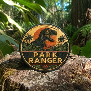 May include: A circular embroidered patch featuring a black silhouette of a Tyrannosaurus Rex against an orange sunset background. The design includes green palm trees and foliage, with the text PARK RANGER in gold letters on a black base.