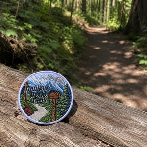 May include: A circular embroidered patch featuring a mountain landscape with a mailbox on a post, a winding trail, and green pine trees. The text "Mailbox Peak Trail" is stitched in dark blue lettering across the center of the design.