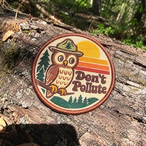 Puede incluir: Un parche bordado circular que muestra un búho marrón con un sombrero de guardabosques verde con un icono de árbol. El diseño incluye un atardecer amarillo y naranja, pinos verde oscuro y el texto marrón Don't Pollute sobre fondo beige.