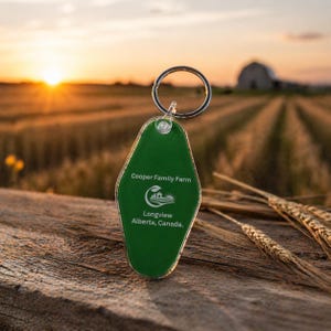 Op de afbeelding: Een groene sleutelhanger met een zilveren ring, met de tekst "Cooper Family Farm" en een logo. De sleutelhanger staat tegen een achtergrond van een zonsondergang boven een tarweveld. De tekst "Longview Alberta, Canada" is ook zichtbaar.