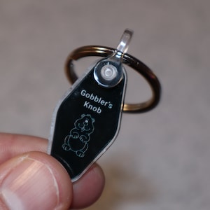 Könnte beinhalten: Ein silberner Schlüsselanhänger mit einem schwarzen Anhänger, auf dem "Gobbler's Knob" steht und ein Cartoon-Bild eines Murmeltiers zu sehen ist.