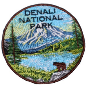 May include: A round, embroidered patch with a brown bear, a lake, and a mountain range. The text "DENALI NATIONAL PARK" is in black lettering.