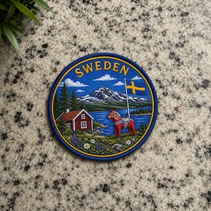 Puede incluir: Un parche bordado circular que muestra un paisaje de Suecia con la palabra SWEDEN en letras amarillas en la parte superior. La escena incluye una casa roja, un caballo de Dalecarlia, una bandera azul y amarilla, un lago, pinos y una montaña.