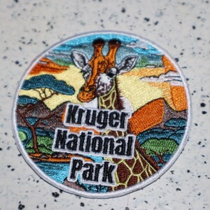 May include: A round embroidered patch featuring a giraffe with a colorful background. The patch says "Kruger National Park" in black text.