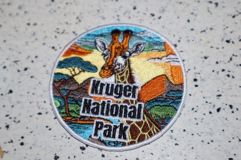 May include: A round embroidered patch featuring a giraffe with a colorful background. The patch says "Kruger National Park" in black text.