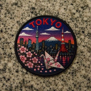 May include: A circular embroidered patch featuring a Tokyo cityscape at sunset with the text TOKYO in red. The design includes the Tokyo Skytree, Tokyo Tower, Mount Fuji, a white origami crane with pink floral patterns, and cherry blossom branches.
