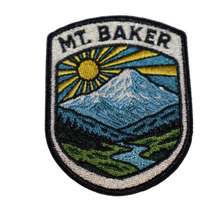 May include: Embroidered patch featuring a mountain landscape with the text "MT. BAKER". The design includes a snow-capped mountain, a sunburst, a river, and trees, all within a shield shape. The patch has a black border.
