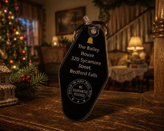May include: A black, teardrop-shaped motel-style keychain with a metal ring. It features a yellow bell icon and white text reading: The Bailey House, 320 Sycamore Street, Bedford Falls, and a circular logo stating: Drop in any mail box, we guarantee postage.