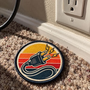 May include: A circular embroidered patch featuring a black electrical plug with yellow prongs and lightning bolts. The background consists of horizontal stripes in yellow, orange, and red, all enclosed within a dark blue stitched border.