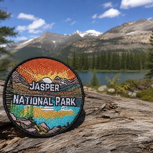 Può includere: Una toppa ricamata circolare che raffigura un paesaggio del Jasper National Park. Il design include un vivace sole nascente arancione e giallo sopra le montagne, un lago blu e pini verdi, con la scritta Jasper National Park in bianco.