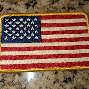 May include: A rectangular embroidered patch featuring the United States flag with a blue canton containing white stars and alternating red and white horizontal stripes, all enclosed by a thick, golden-yellow stitched border.