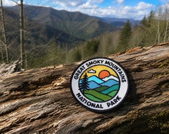 May include: A circular embroidered patch featuring a landscape design of the Great Smoky Mountains National Park. The patch displays green rolling hills, blue mountain peaks, a yellow sky, and an orange sun, with black text around the border.
