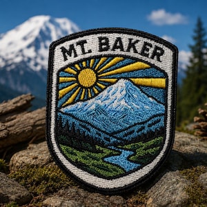 May include: Embroidered patch featuring a mountain landscape with the text "MT. BAKER". The design includes a snow-capped mountain, a sunburst, a river, and trees, all in shades of blue, green, and yellow. The patch is bordered in black and white.