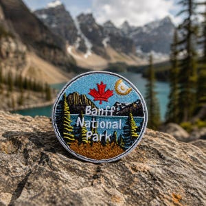 May include: Embroidered patch featuring a scenic design of Banff National Park. The patch includes a red maple leaf, a crescent moon, mountains, and pine trees. The text "Banff National Park" is displayed on the patch.