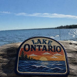 Pode incluir: Um emblema bordado semicircular com o texto LAKE ONTARIO em letras azul-escuras. O design retrata um pôr do sol sobre a água com um sol amarelo, céu laranja, nuvens brancas e ondas azul-escuras, emoldurado por uma borda azul-escura.