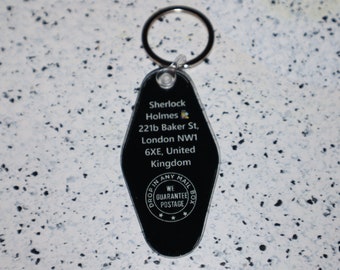 Pode incluir: Chaveiro preto com o texto "Sherlock Holmes, 221b Baker St, London NW1 6XE, United Kingdom." O chaveiro tem um anel de metal e uma alça de plástico transparente.