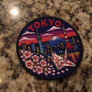 Puede incluir: Un parche bordado circular que muestra un vibrante paisaje urbano de Tokio al atardecer. El diseño incluye la palabra TOKYO en rojo, el Tokyo Skytree, la Torre de Tokio, el Monte Fuji, una grulla de origami blanca con flores y cerezos en flor.