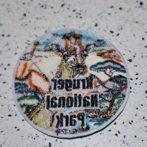 May include: A round, white patch with colorful embroidered details. The patch features a landscape scene with trees, a building, and the text "1998 National Park".