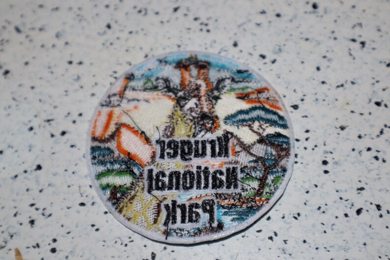 May include: A round, white patch with colorful embroidered details. The patch features a landscape scene with trees, a building, and the text "1998 National Park".