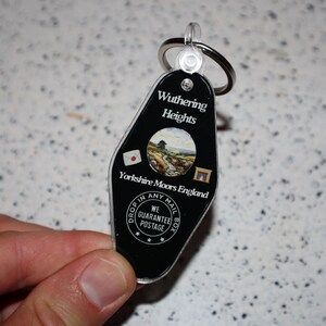 May include: Black plastic keychain with a silver key ring. The keychain has the words "Wuthering Heights" and "Yorkshire Moors England" printed on it. A small circular image of a landscape is in the center. The keychain also has the text "Drop in any mail box" and "We guarantee postage".