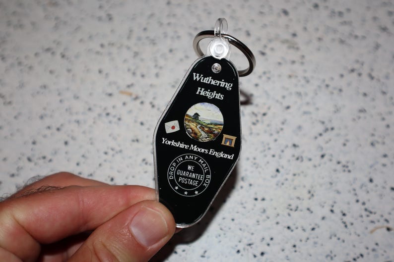 May include: Black plastic keychain with a silver key ring. The keychain has the words "Wuthering Heights" and "Yorkshire Moors England" printed on it. A small circular image of a landscape is in the center. The keychain also has the text "Drop in any mail box" and "We guarantee postage".
